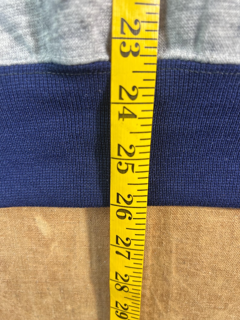 May include: Close-up of a gray and navy blue fabric with a yellow measuring tape. The tape shows measurements from 23 to 29 inches. The fabric appears to be a section of a garment.