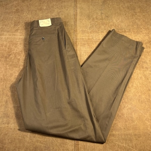 Puede incluir: Un pantalón de color caqui, doblado y colocado plano. El pantalón tiene cierre de botón, bolsillos y trabillas para el cinturón. Una etiqueta está adherida a la cintura. La tela parece ser de algodón o mezcla de peso medio.