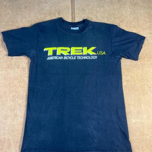 Puede incluir: Camiseta azul marino oscuro de manga corta con la palabra "TREK" en verde neón y la palabra "USA" en verde neón. Debajo, el texto "AMERICAN BICYCLE TECHNOLOGY".