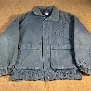 May include: A gray work jacket with a button-up front, two chest pockets, and two lower pockets. The jacket has a collared neckline and elastic cuffs and waistband. The jacket is made of a durable material.