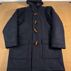 May include: A dark navy blue duffle coat featuring a hood, long sleeves, and two large front patch pockets. The coat is secured with four brown wooden toggle closures attached to black rope loops, providing a classic front-fastening design.