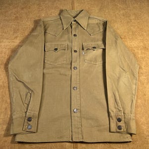 May include: A tan button-down shirt with two chest pockets and buttoned cuffs. The shirt has a pointed collar and a Western-style yoke. The buttons are silver-colored. The shirt is made of a durable fabric.