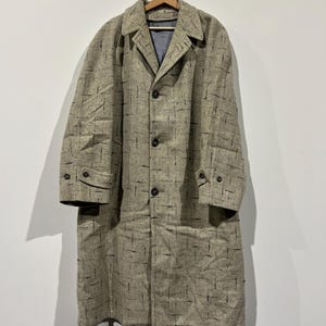 May include: A beige overcoat with a black grid pattern, hanging on a wooden hanger. The coat features a notched collar, button fastenings, and two front pockets. The sleeves have buttoned cuffs.