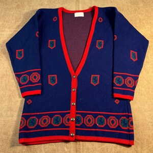 May include: A navy blue V-neck cardigan sweater featuring a bright red trim along the button placket, cuffs, and hem. The knit garment is decorated with repeating red and green shield-shaped motifs and geometric circular patterns throughout.