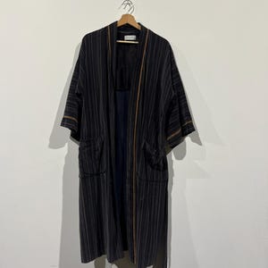 Op de afbeelding: Een lange, openstaande kimono met verticale strepen in zwart- en grijstinten, hangend aan een houten hanger. De kimono heeft twee voorzakken en bruine biezen langs de randen en mouwen. De kimono is ongeveer 140 cm lang.