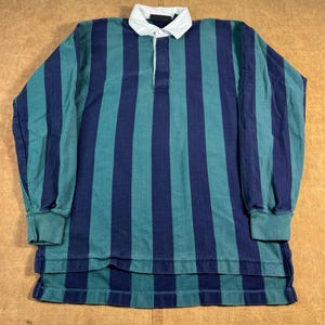 May include: A long-sleeved rugby shirt with vertical stripes in teal and navy blue. The shirt has a white collar and a button-down placket. The cuffs and hem are solid teal.