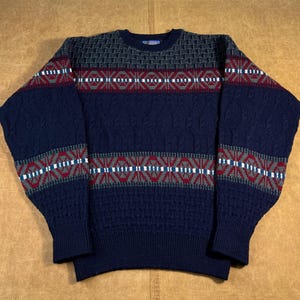 Könnte beinhalten: Ein marineblauer Pullover mit Rundhalsausschnitt und Muster. Der Pullover hat einen strukturierten Strick mit geometrischen Mustern in Rot, Weiß und Grau. Die Ärmel und die obere Brust haben ein sich wiederholendes Muster.
