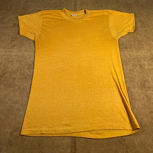 May include: Mustard yellow short-sleeved t-shirt. The shirt has a crew neck and a slightly faded appearance. The fabric appears to be a lightweight material, suitable for casual wear. The shirt is laid flat on a brown surface.