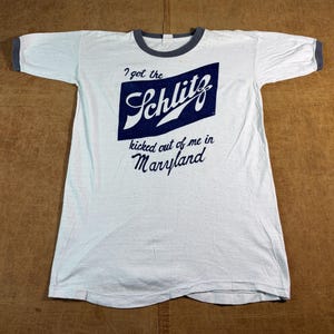 May include: A white ringer t-shirt with dark gray trim on the collar and sleeve cuffs. The front features a dark blue rectangular graphic with the white text Schlitz and the black printed text I got the kicked out of me in Maryland.