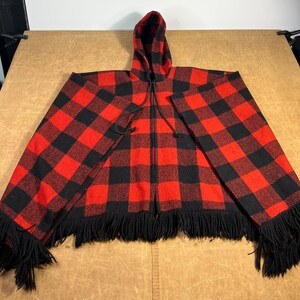 May include: A red and black plaid hooded cape with a full-length zipper. The cape features a black fringe along the bottom edge and is made of a thick, warm material. The hood is lined with the same plaid pattern.