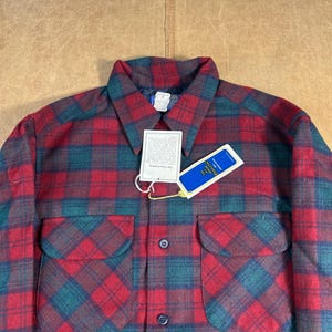 May include: A red and green checked shirt with two front pockets. The shirt has a collar and a tag attached. The checked pattern is a classic design, and the shirt appears to be made of a warm, durable material.