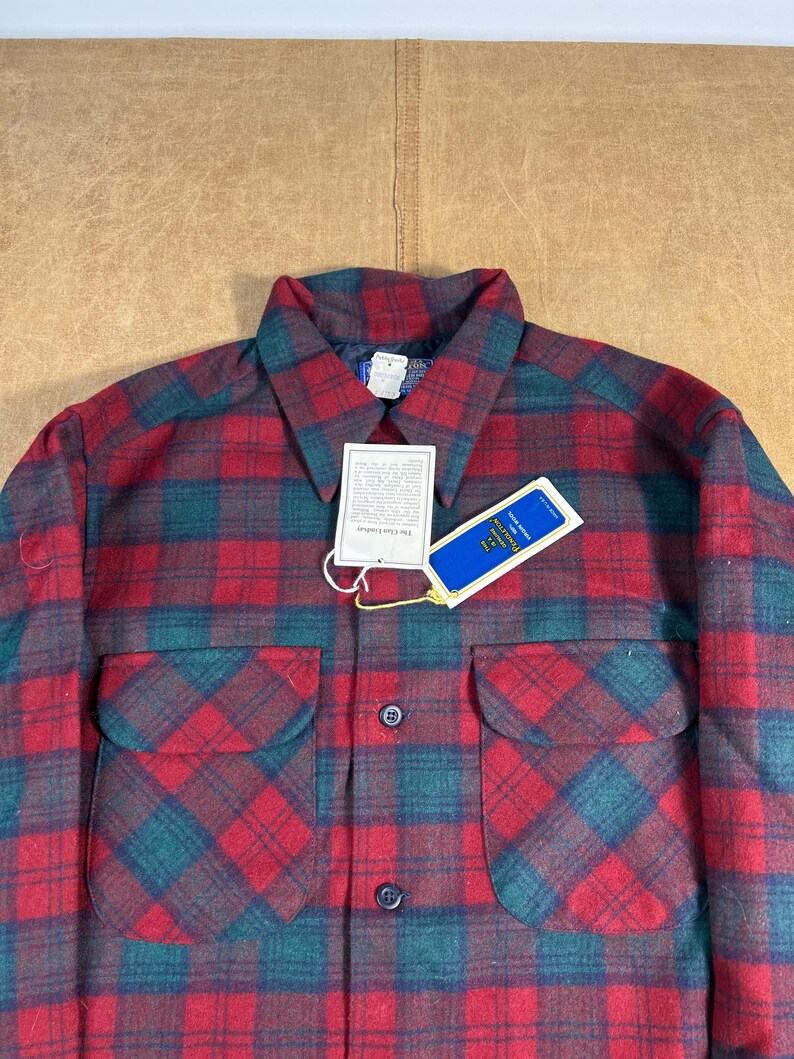May include: A red and green checked shirt with two front pockets. The shirt has a collar and a tag attached. The checked pattern is a classic design, and the shirt appears to be made of a warm, durable material.