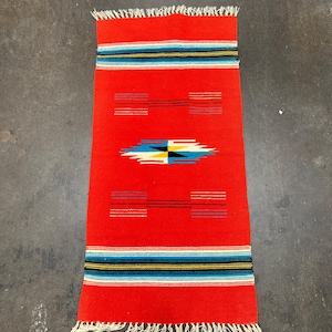 Op de afbeelding: Een rechthoekig, rood geweven vloerkleed met een geometrisch ontwerp. Het vloerkleed heeft horizontale strepen in blauw, zwart en wit nabij de boven- en onderranden. Een centraal ontwerp omvat een ruitvorm met blauwe, gele en zwarte elementen. Het vloerkleed heeft franjes.