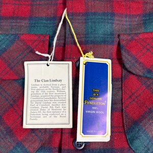 May include: Close-up of a red, green, and blue plaid Pendleton shirt. The shirt has two tags attached. One tag reads "The Clan Lindsay" and the other reads "This is a Genuine Pendleton 100% Virgin Wool Made in USA."
