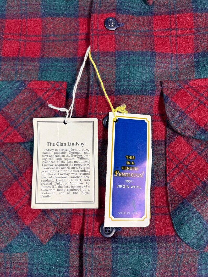 May include: Close-up of a red, green, and blue plaid Pendleton shirt. The shirt has two tags attached. One tag reads "The Clan Lindsay" and the other reads "This is a Genuine Pendleton 100% Virgin Wool Made in USA."