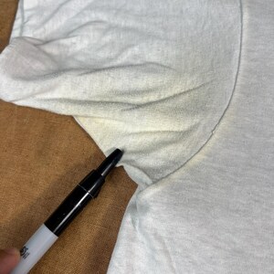 May include: Close-up of a white t-shirt with a black marker pointing at the armpit area. The shirt shows signs of wear and discoloration, with visible stains. The fabric appears to be a lightweight cotton.