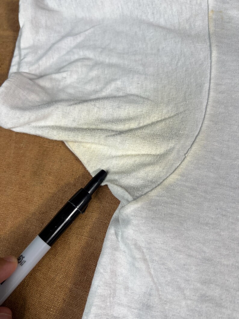May include: Close-up of a white t-shirt with a black marker pointing at the armpit area. The shirt shows signs of wear and discoloration, with visible stains. The fabric appears to be a lightweight cotton.