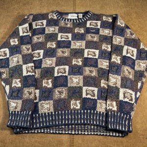 May include: A blue, brown, and white checkered pattern sweater. The sweater has a ribbed crew neck, cuffs, and hem. The pattern is made up of squares in various shades of blue, brown, and white.