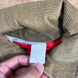May include: Close-up of a tan corduroy jacket collar with a red lining. A white tag with text is visible. The jacket appears to be vintage.