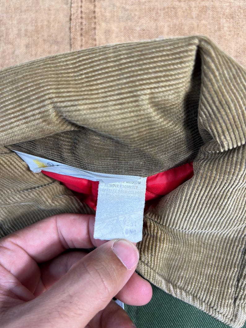 May include: Close-up of a tan corduroy jacket collar with a red lining. A white tag with text is visible. The jacket appears to be vintage.