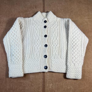 May include: Cream-colored cable-knit cardigan sweater with a high collar and dark buttons. The sweater features intricate cable and textured knit patterns. A classic, warm garment for cooler weather.