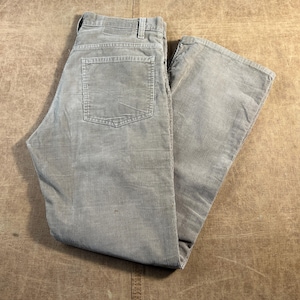 Könnte beinhalten: Eine hellgraue Cordhose mit klassischem Fünf-Taschen-Design, Knopf- und Reißverschluss, Gürtelschlaufen am Bund und einer aufgesetzten Tasche auf der Rückseite. Der Stoff weist durchgehend eine vertikal gerippte Textur auf.
