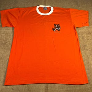 Könnte beinhalten: Oranges T-Shirt mit weißem Kragen und kurzen Ärmeln. Das Shirt zeigt eine kleine Grafik von zwei Zeichentigern mit dem Text "Tiger Coffee Shop" auf der Vorderseite. Das Shirt ist aus einem weichen Material gefertigt.