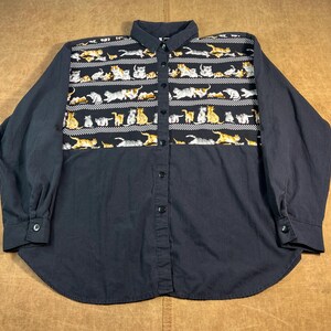 May include: A dark navy blue button-down shirt with long sleeves. The upper chest area features a repeating pattern of various cat illustrations in shades of yellow, white, and gray. The shirt has a classic collar and buttoned cuffs.