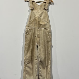May include: Faded tan overalls hanging on a wooden hanger. The dungarees feature adjustable straps, a bib pocket, and multiple pockets on the legs. The fabric shows signs of wear and tear, with visible stitching. The garment is displayed against a plain white wall.