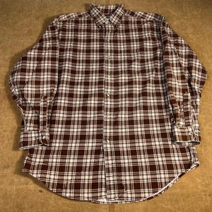 May include: A brown and white checked button-down shirt with long sleeves. The shirt features a classic collar and buttoned cuffs. The fabric has a woven texture. The shirt is designed with a curved hemline.
