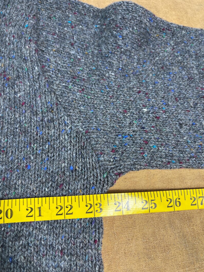 May include: Close-up of a gray knit sweater with flecks of blue, red, and green. A yellow measuring tape shows a length of 20 to 27 inches. The sweater has a textured appearance.