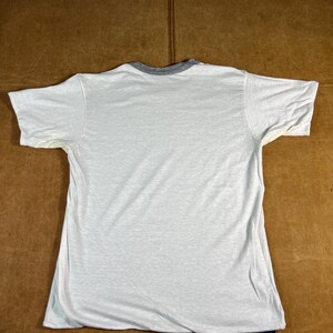 May include: A white t-shirt with a gray collar and short sleeves. The shirt is laid flat on a brown surface. There is a small blue tag at the bottom of the shirt.