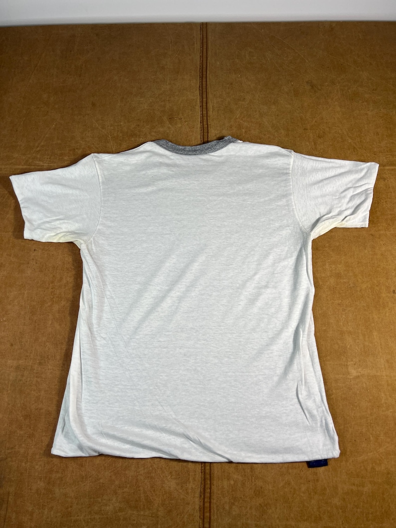 May include: A white t-shirt with a gray collar and short sleeves. The shirt is laid flat on a brown surface. There is a small blue tag at the bottom of the shirt.