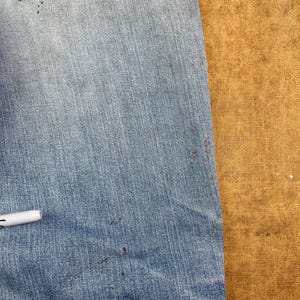 May include: Close-up of light blue denim jeans with a distressed look. The jeans have a worn pocket with a yellowed patch and several dark spots. A white pen rests on the denim.