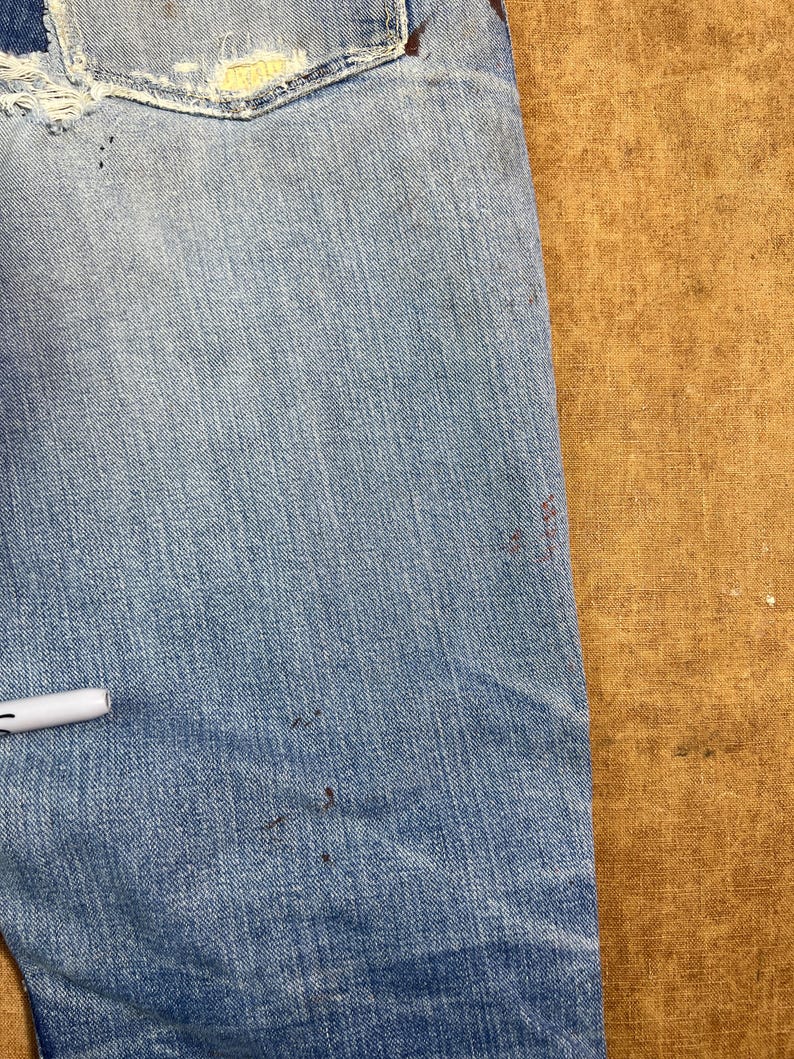May include: Close-up of light blue denim jeans with a distressed look. The jeans have a worn pocket with a yellowed patch and several dark spots. A white pen rests on the denim.