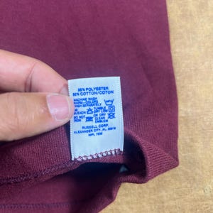 May include: Close-up of a maroon-colored garment with a white tag. The tag reads "50% POLYESTER 50% COTTON/COTON" and includes washing instructions. The garment's label also indicates "RUSSELL CORP ALEXANDER CITY, AL 35018 WPL 7832".