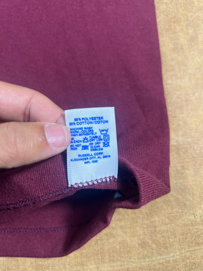 May include: Close-up of a maroon-colored garment with a white tag. The tag reads "50% POLYESTER 50% COTTON/COTON" and includes washing instructions. The garment's label also indicates "RUSSELL CORP ALEXANDER CITY, AL 35018 WPL 7832".