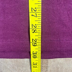 May include: Close-up of a deep purple fabric with a yellow measuring tape. The tape shows measurements from 26 to 33 inches. The fabric appears to be a t-shirt or similar garment.