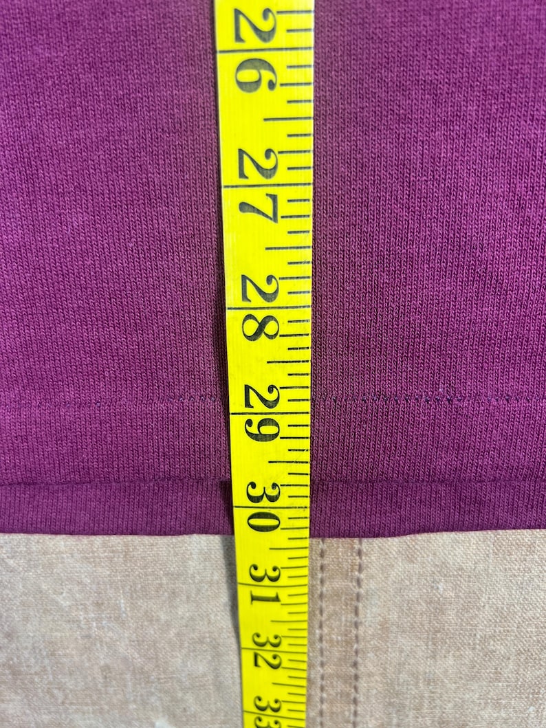 May include: Close-up of a deep purple fabric with a yellow measuring tape. The tape shows measurements from 26 to 33 inches. The fabric appears to be a t-shirt or similar garment.