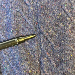 May include: Close-up of a textured, dark blue fabric with flecks of red, yellow, and green. A black pen is in the lower left corner, pointing towards the fabric. The fabric appears to be a knit or woven material.