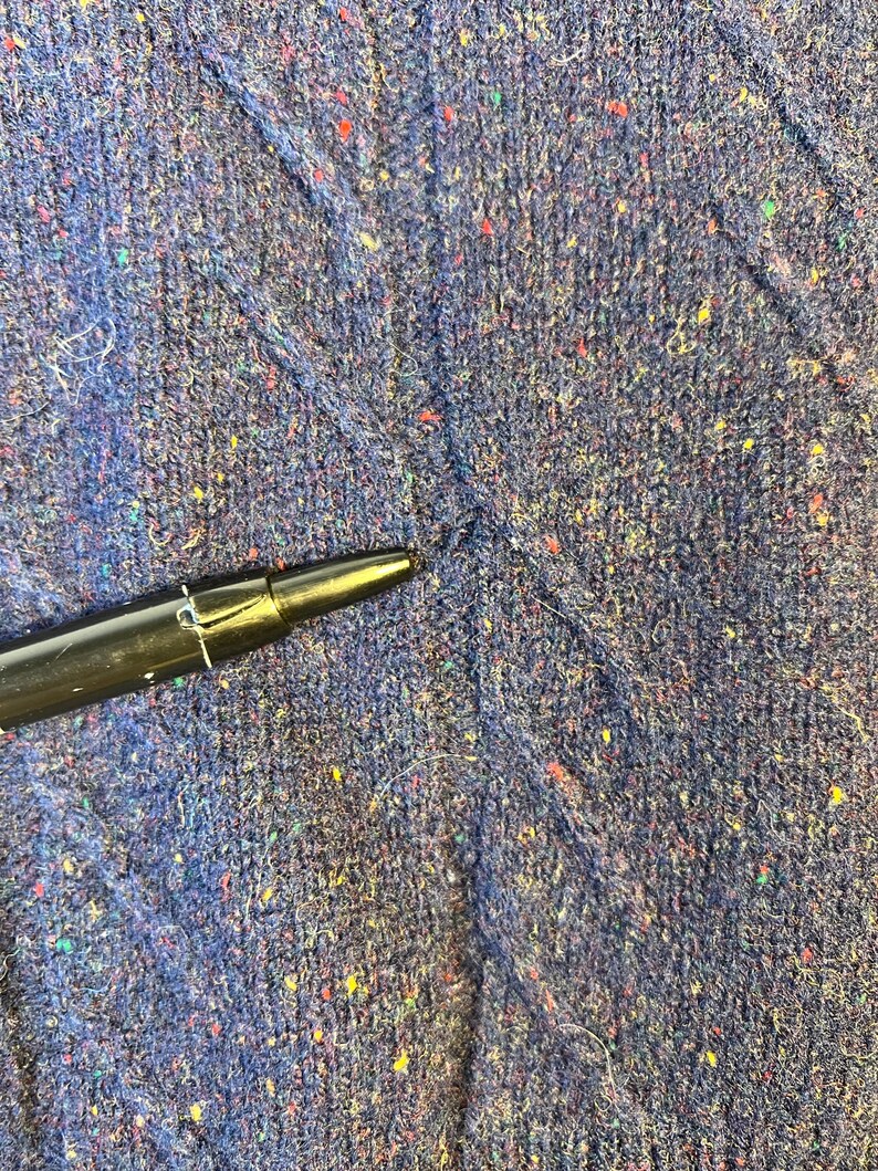 May include: Close-up of a textured, dark blue fabric with flecks of red, yellow, and green. A black pen is in the lower left corner, pointing towards the fabric. The fabric appears to be a knit or woven material.