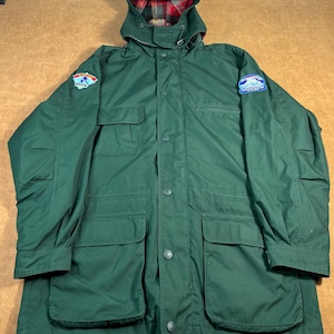 Op de afbeelding: Een donkergroene jas met capuchon, meerdere zakken en een knoopsluiting. De voering van de capuchon heeft een rood-zwart ruitpatroon. Op de mouwen zijn patches zichtbaar. De jas is ontworpen voor buitenactiviteiten.