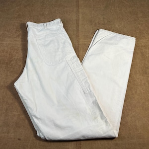 May include: Off-white work pants with multiple pockets and a straight leg. The pants are made of a durable fabric and are laid flat on a brown surface. The pants have a classic, casual style.