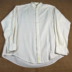 Könnte beinhalten: Cremefarbenes Langarmhemd mit Stehkragen und geknöpften Manschetten. Das Hemd hat ein vertikales strukturiertes Muster und eine Knopfleiste vorne. Der Kragen hat einen einzelnen schwarzen Knopf.