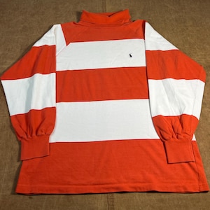 May include: An orange and white striped long-sleeved top with a turtleneck collar. The shirt has a small black logo on the chest. The sleeves are white with orange cuffs. A classic, casual top.