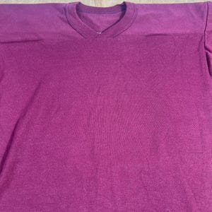 May include: A solid magenta-colored V-neck t-shirt. The shirt has a simple design with a ribbed neckline and shoulder seams. The fabric appears to be a soft knit material.
