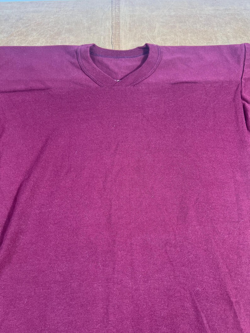 May include: A solid magenta-colored V-neck t-shirt. The shirt has a simple design with a ribbed neckline and shoulder seams. The fabric appears to be a soft knit material.