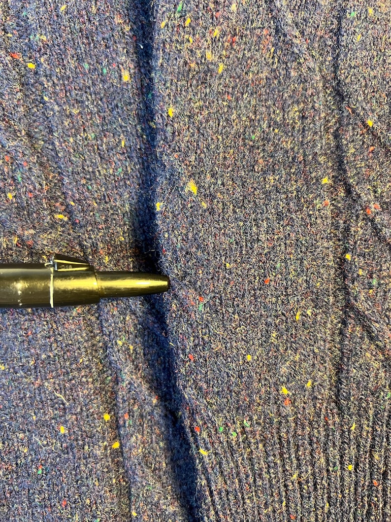 May include: Close-up of a navy blue knit fabric with flecks of yellow, red, and green. The fabric has a textured appearance and is likely from a sweater or other garment. A black pen is partially visible.