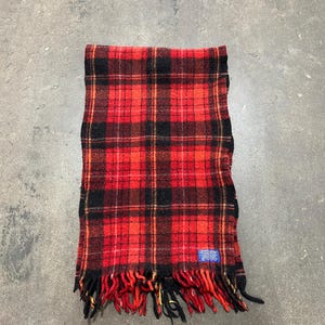 May include: A red, black, and gold plaid scarf with fringed edges. The scarf has a blue tag on the bottom right corner. The scarf is made of a woven material.