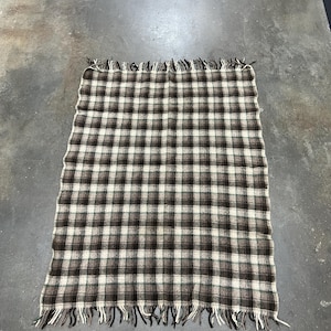 Puede incluir: Una manta rectangular con un patrón de cuadros marrón, blanco y negro. La manta tiene flecos en los bordes. La manta está sobre una superficie gris.